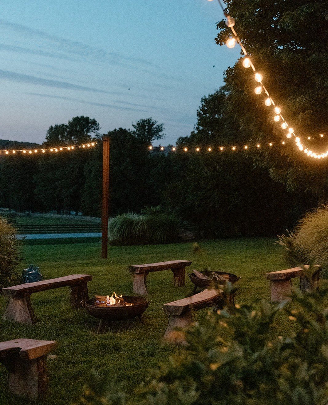 35 Backyard Wedding Reception Ideas for an Unforgettable Day 28 Metal fire pit with burning flames surrounded by wooden benches under string lights.