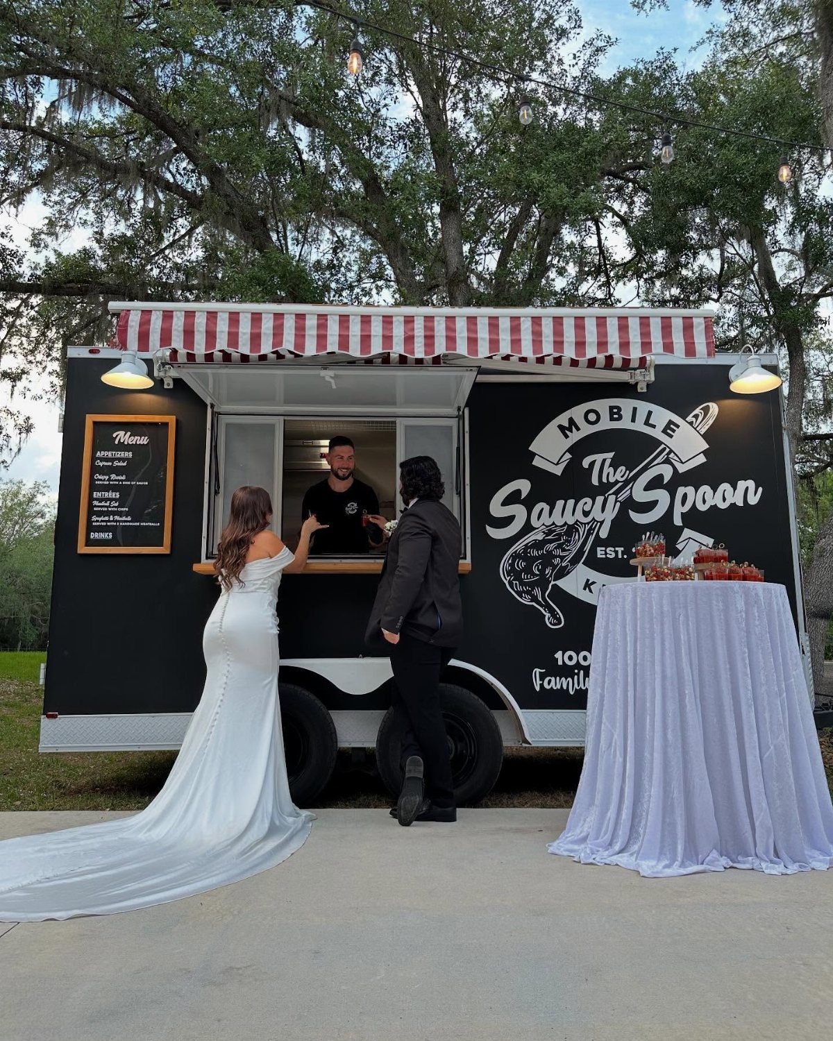 35 Backyard Wedding Reception Ideas for an Unforgettable Day 4 Black food truck with red striped awning serving a bride and groom outdoors.