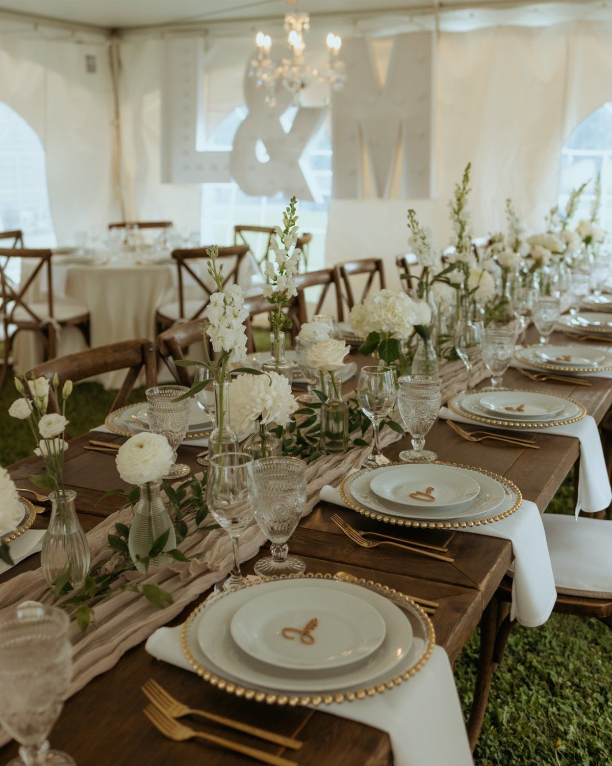 35 Backyard Wedding Reception Ideas for an Unforgettable Day 14 Long wooden banquet table with greenery and white floral centerpieces under a wedding tent.