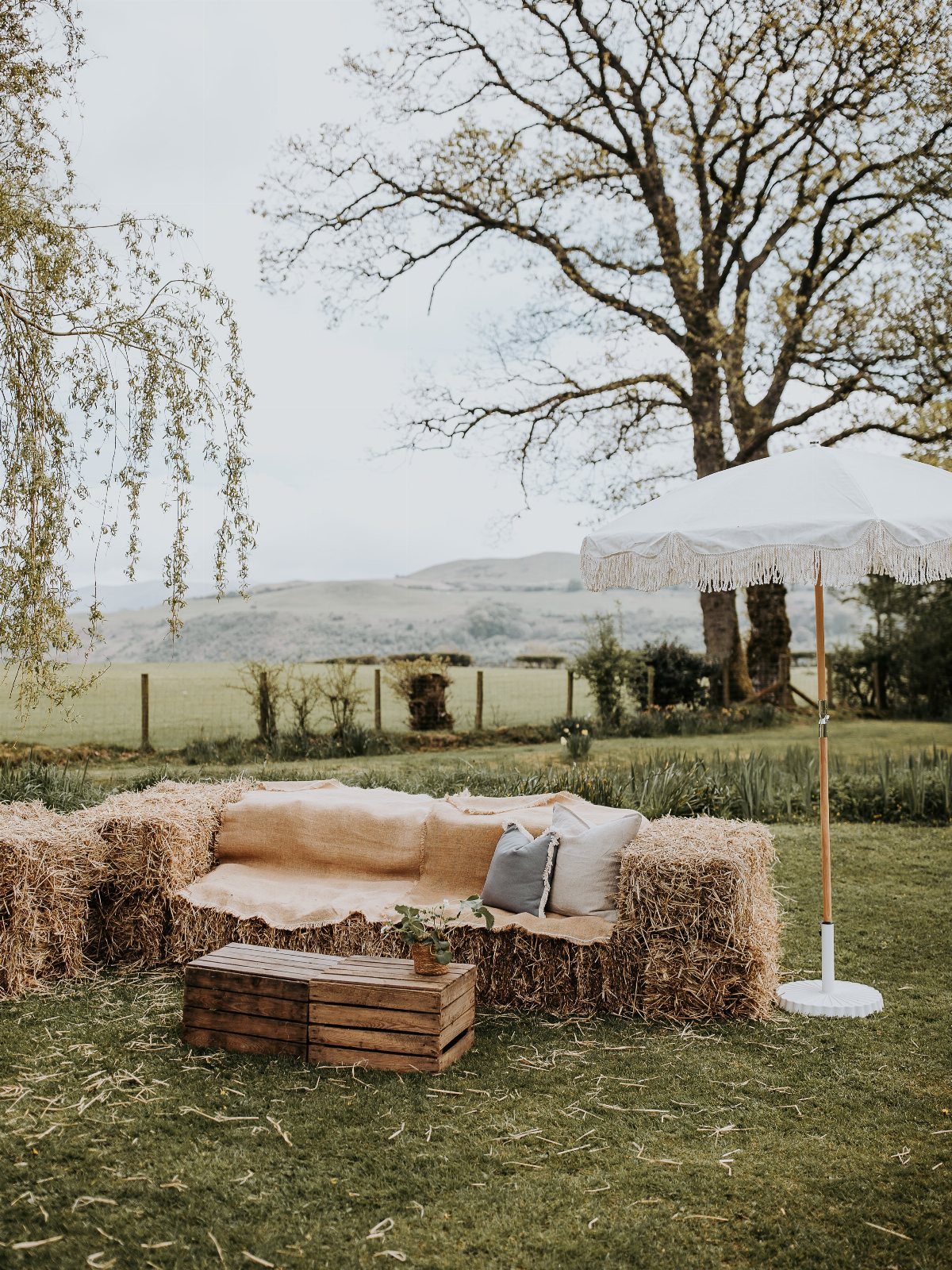 35 Backyard Wedding Reception Ideas for an Unforgettable Day 23 Hay bale seating with blankets and pillows under a white fringed umbrella outdoors.