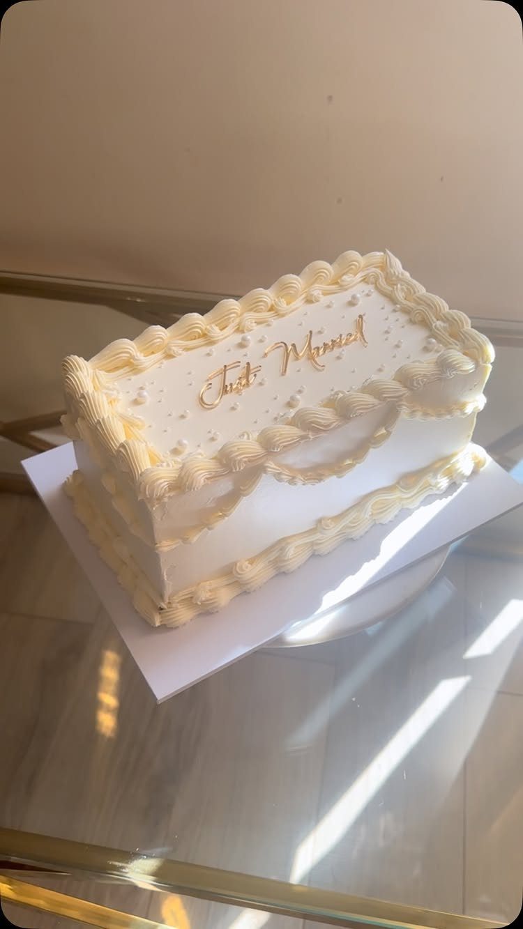 5 Wedding Cake Just Married Ideas for Real Budgets 5 A white rectangular cake with gold Just Married text and decorative frosting borders on a glass table.