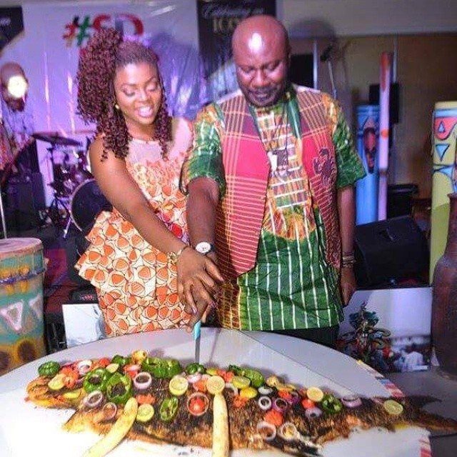 Couple in traditional African clothing cutting a large garnished grilled fish together.