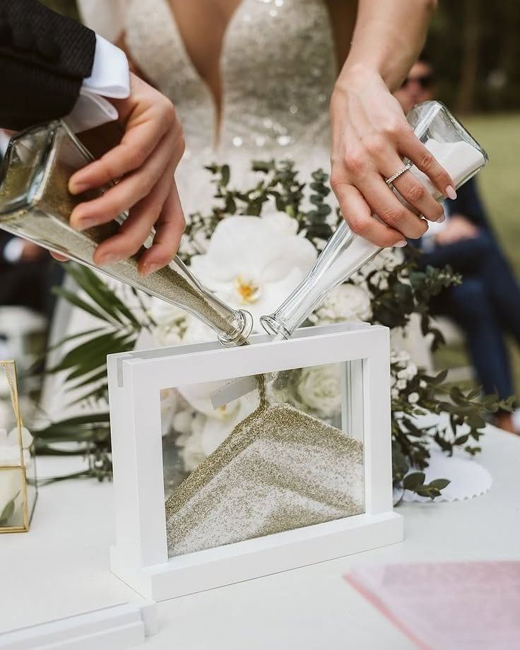 Couple pouring gold and white sand into a white framed glass vessel