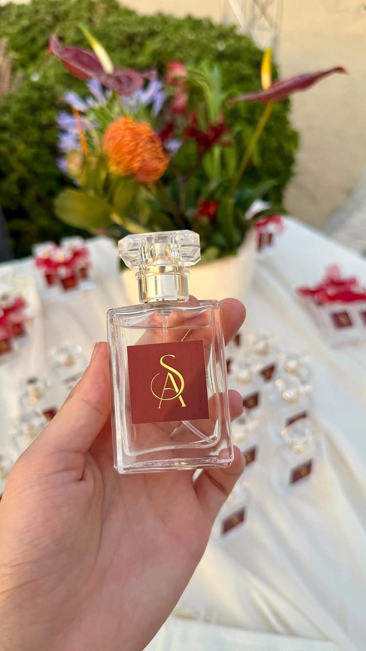 Wedding Detail Shots: 21 Ideas for Your Photographer 17 A hand holding a small clear glass perfume bottle with a gold monogrammed label against a floral background.