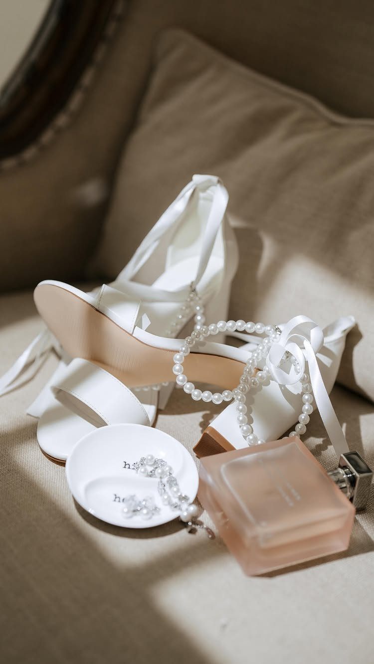 Wedding Detail Shots: 21 Ideas for Your Photographer 14 White satin bridal sandals, pearls, and a perfume bottle arranged on a beige fabric surface.