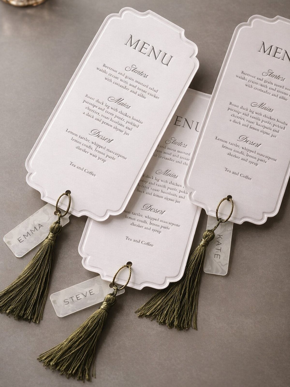 Wedding Detail Shots: 21 Ideas for Your Photographer 16 White scalloped wedding menus with olive green tassels and personalized name tags on a grey background.