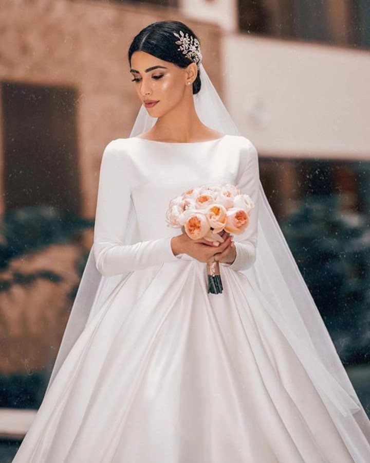 Woman in a white satin A-line wedding dress holding peach roses bouquet.
