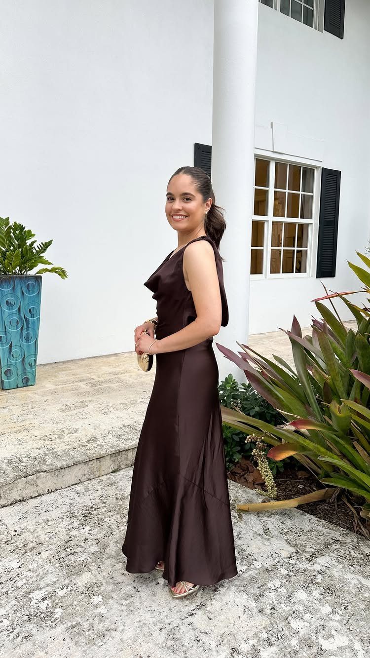 Woman in a dark brown asymmetrical satin dress standing outdoors by a white building.