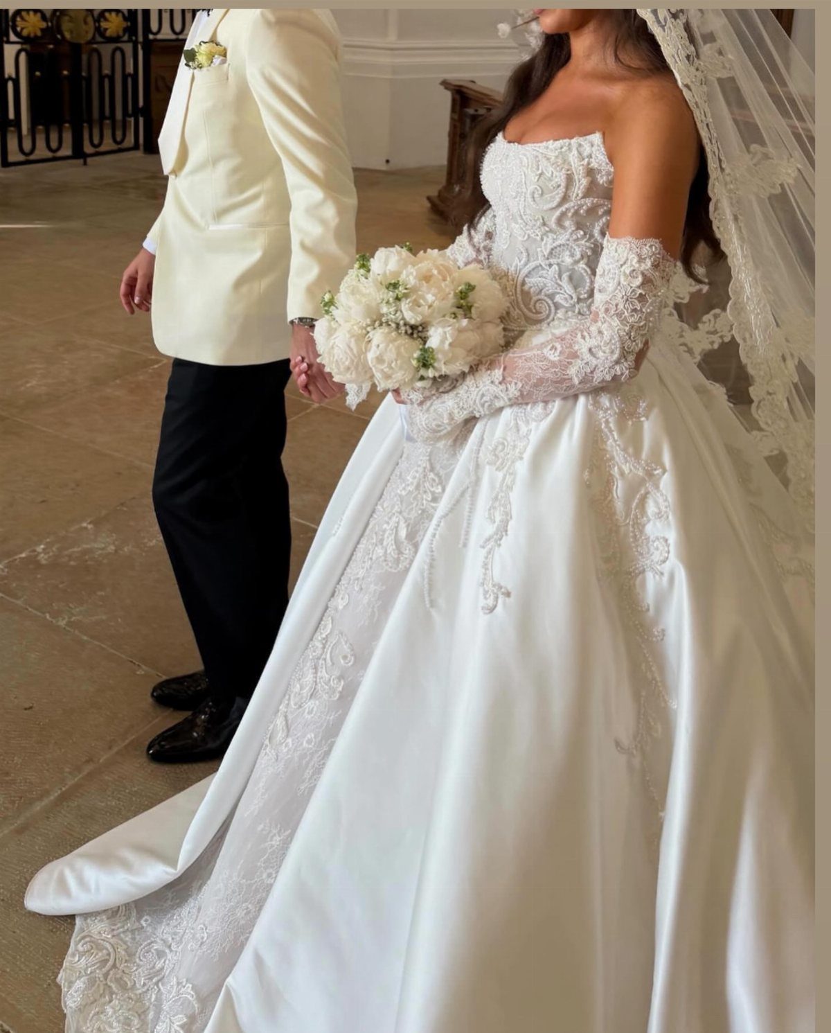 Bride in a shimmering lace and satin ballgown with detachable sleeves holding white flowers.