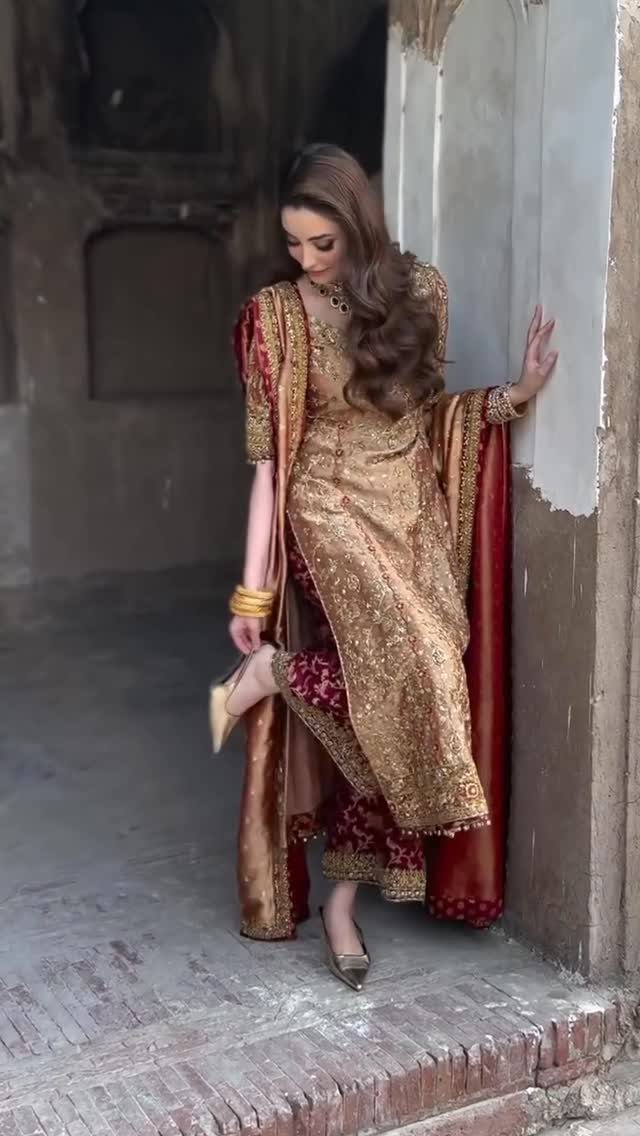 Woman in gold and maroon traditional bridal dress posing against a stone wall.