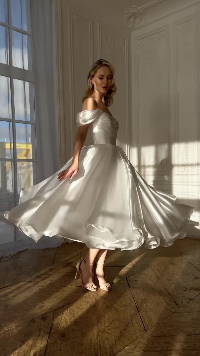Woman in a white off-the-shoulder tea-length satin wedding dress posing in a bright room.