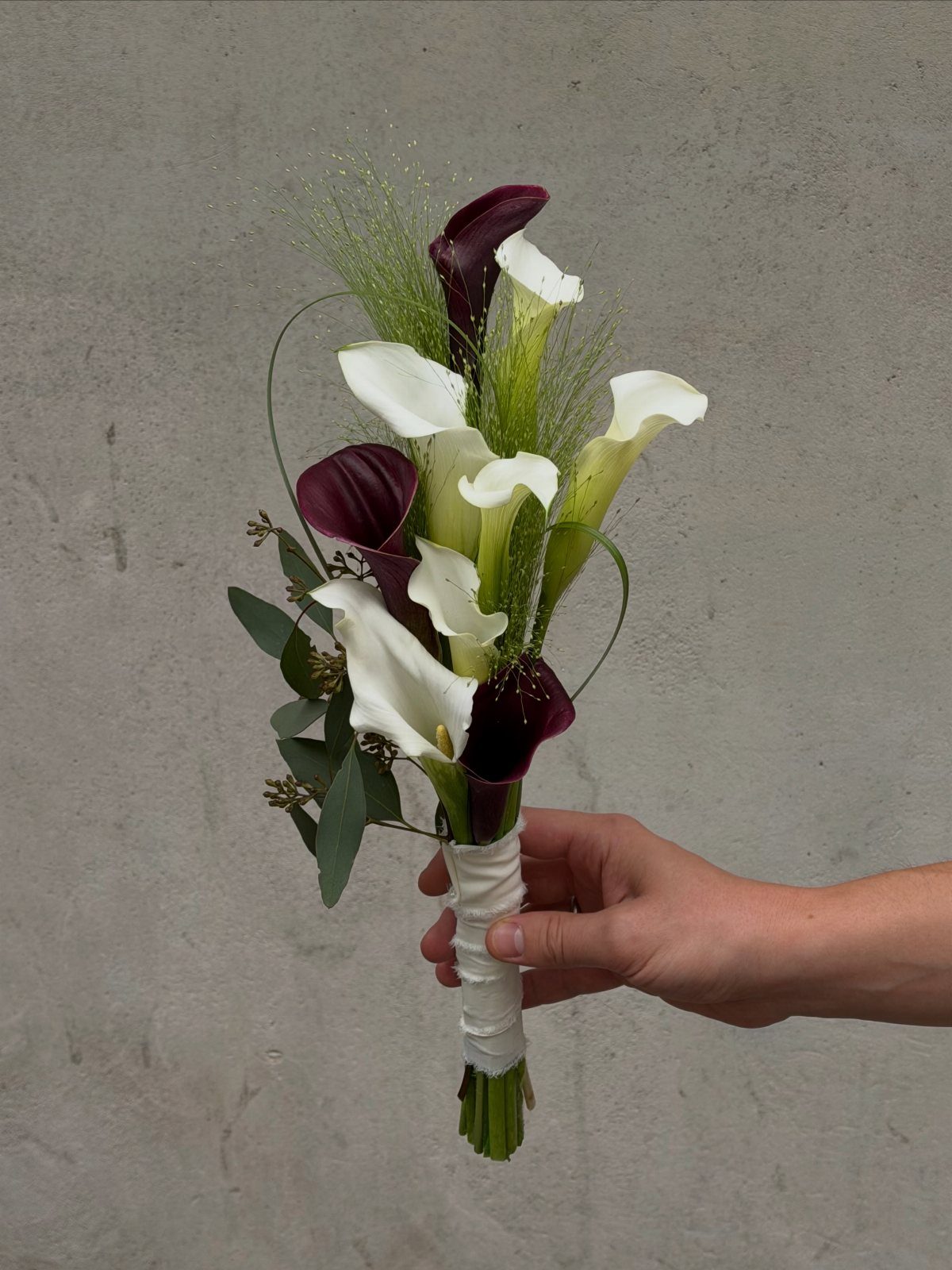 Wedding Flower Trends: 36 Ideas from Jewel Tones to Arches 20 Calla lily bouquet with white and burgundy flowers held against a grey wall.
