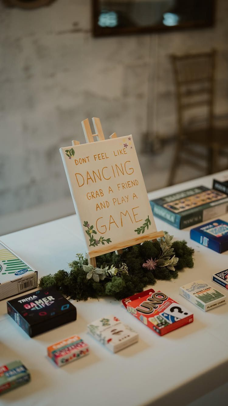 13 Wedding Games Table Ideas Your Guests Will Actually Play 10 A white sign on an easel surrounded by various board games and playing cards on a table.