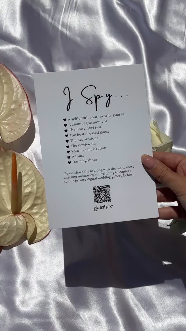 13 Wedding Games Table Ideas Your Guests Will Actually Play 6 A hand holding an I Spy wedding scavenger hunt card over white satin fabric.