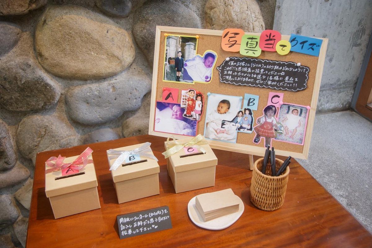 13 Wedding Games Table Ideas Your Guests Will Actually Play 8 A photo quiz setup on a wooden table featuring a corkboard with pictures and labeled ballot boxes.