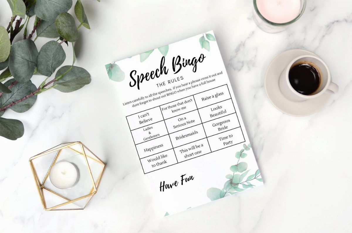 13 Wedding Games Table Ideas Your Guests Will Actually Play 4 Speech Bingo card with eucalyptus leaves on a white marble tabletop next to coffee.