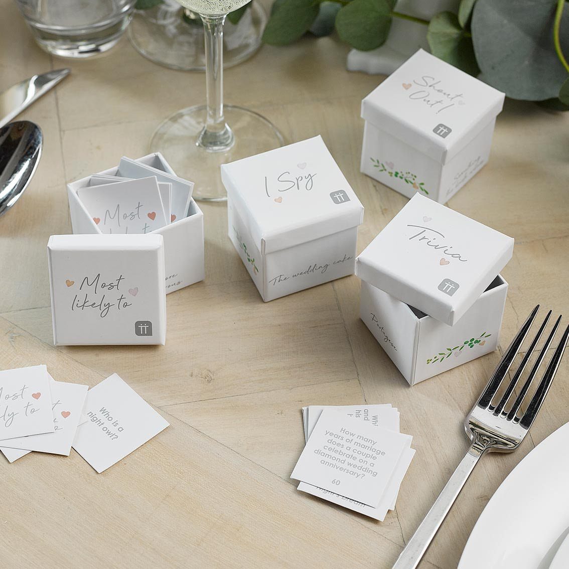 13 Wedding Games Table Ideas Your Guests Will Actually Play 14 Small white boxes containing wedding games like trivia and I Spy scattered on a wooden table.
