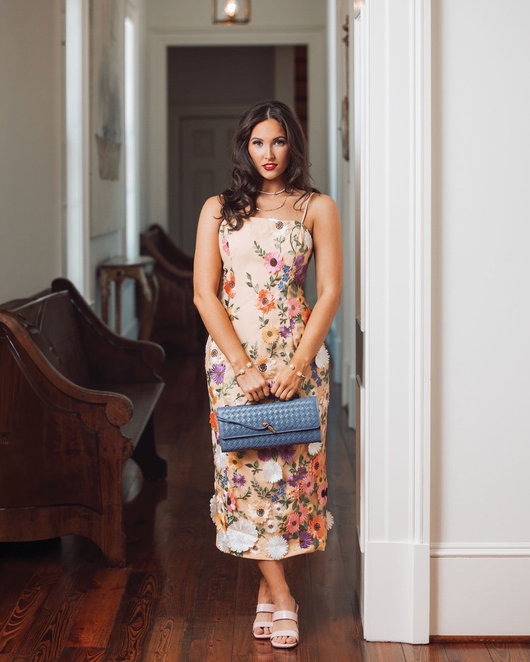 19 Wedding Guest Dress Ideas for Every Season and Style 14 Woman in a cream midi dress with colorful floral embroidery holding a blue clutch.