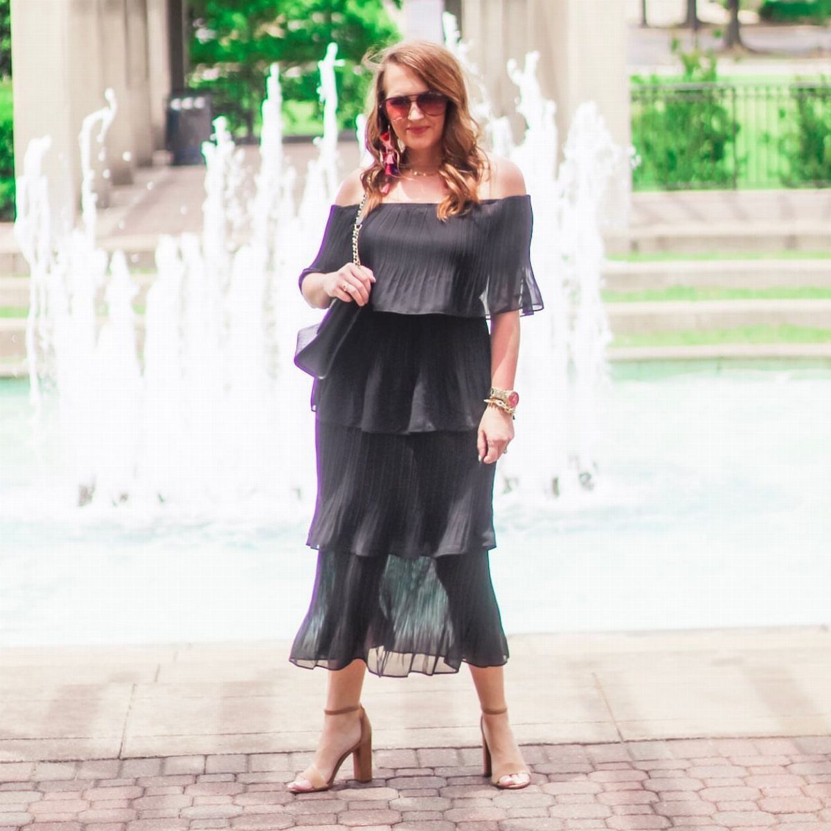 19 Wedding Guest Dress Ideas for Every Season and Style 18 Woman wearing a black off-the-shoulder tiered midi dress posing by a water fountain