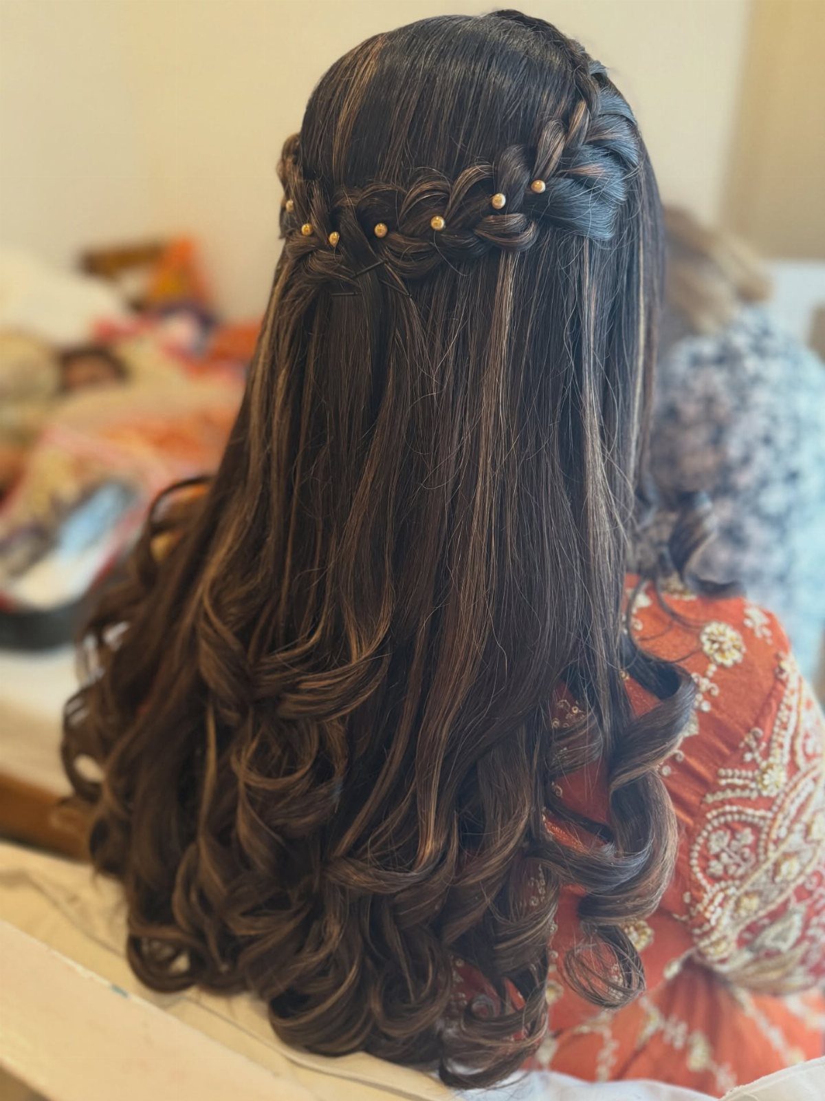 Long dark curly hair styled in a braided half-updo with gold bead accents.