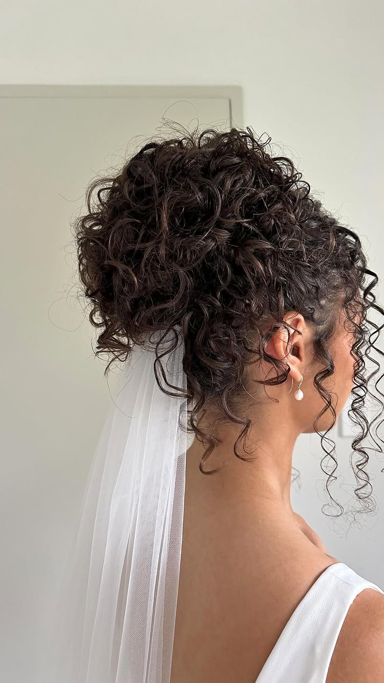 Curly dark hair styled in a high piled updo with a white bridal veil.