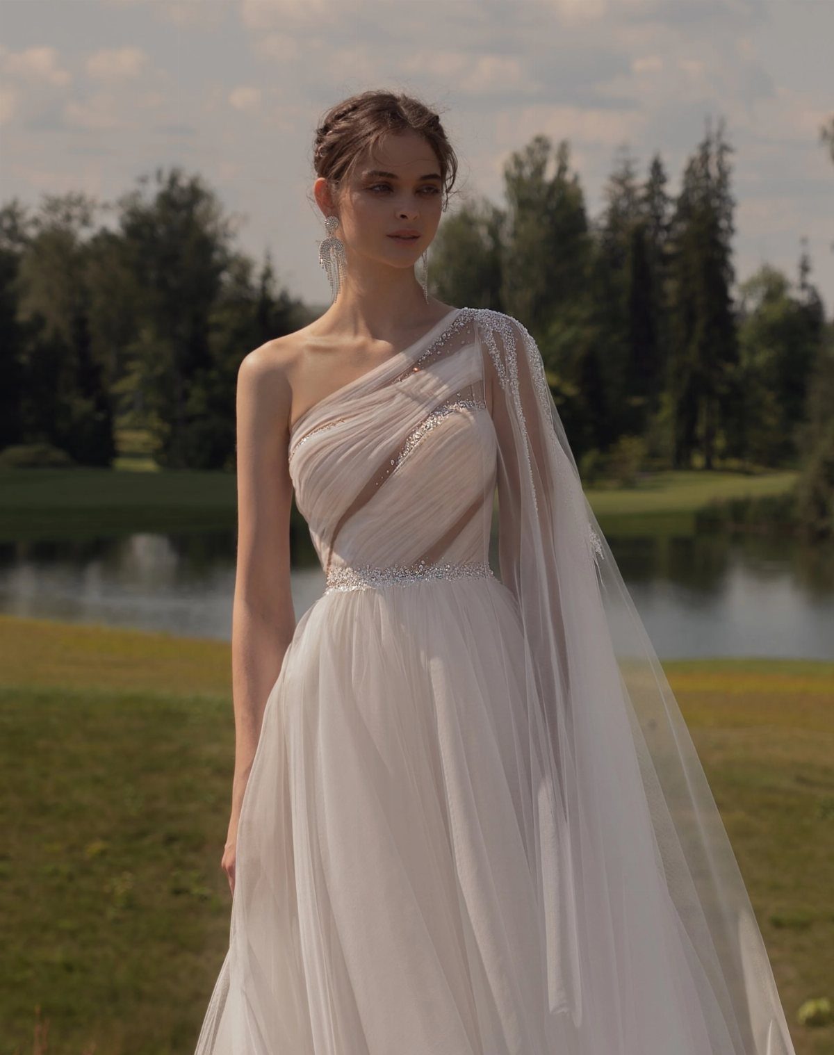 Wedding Outfit Outdoor: 9 Guest Looks for Every Setting 4 A woman in a white one-shoulder asymmetric gown standing in a grassy field near a lake.