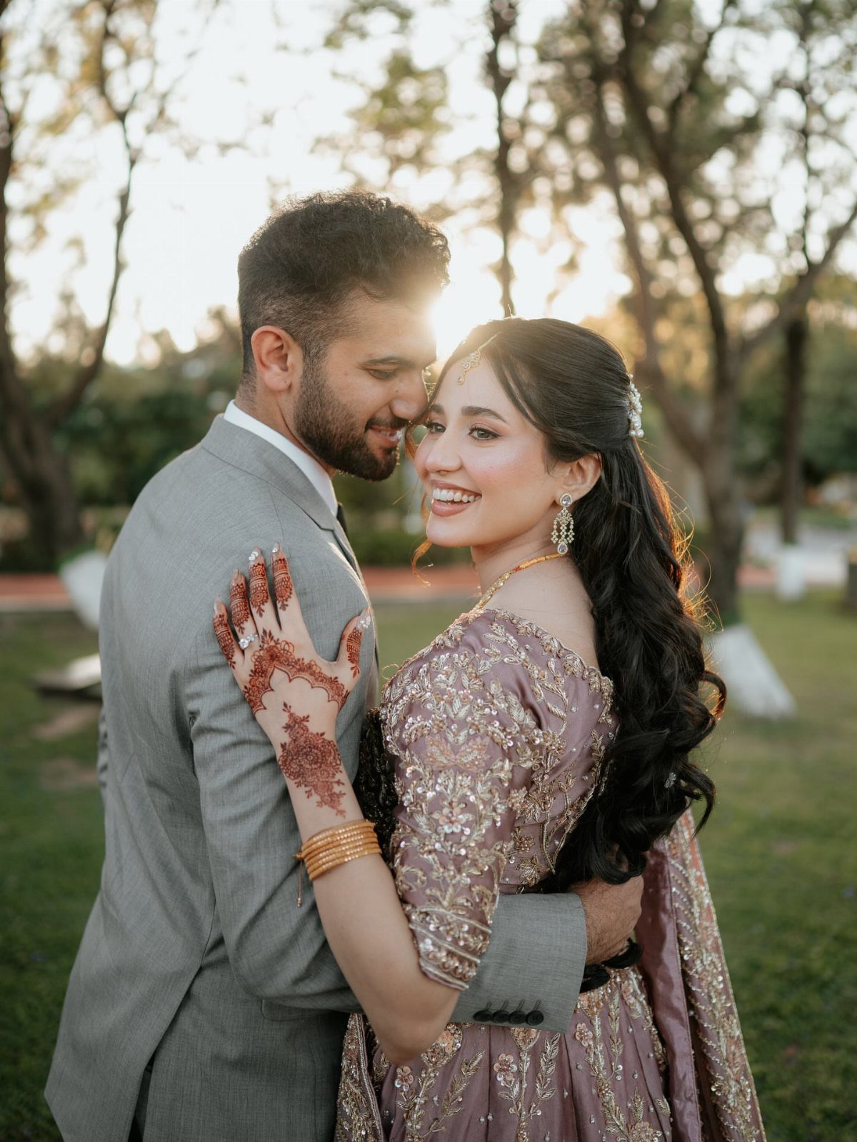 A smiling couple posing outdoors, the woman wearing an embellished mauve lehenga and henna.