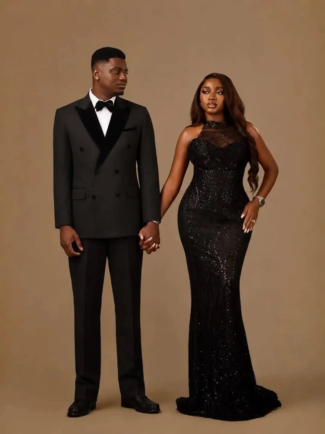 A man in a black tuxedo and a woman in a black gown holding hands.
