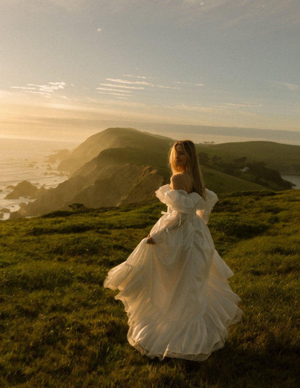 A woman in a white wedding dress standing on a grassy cliff overlooking the ocean at sunset.
