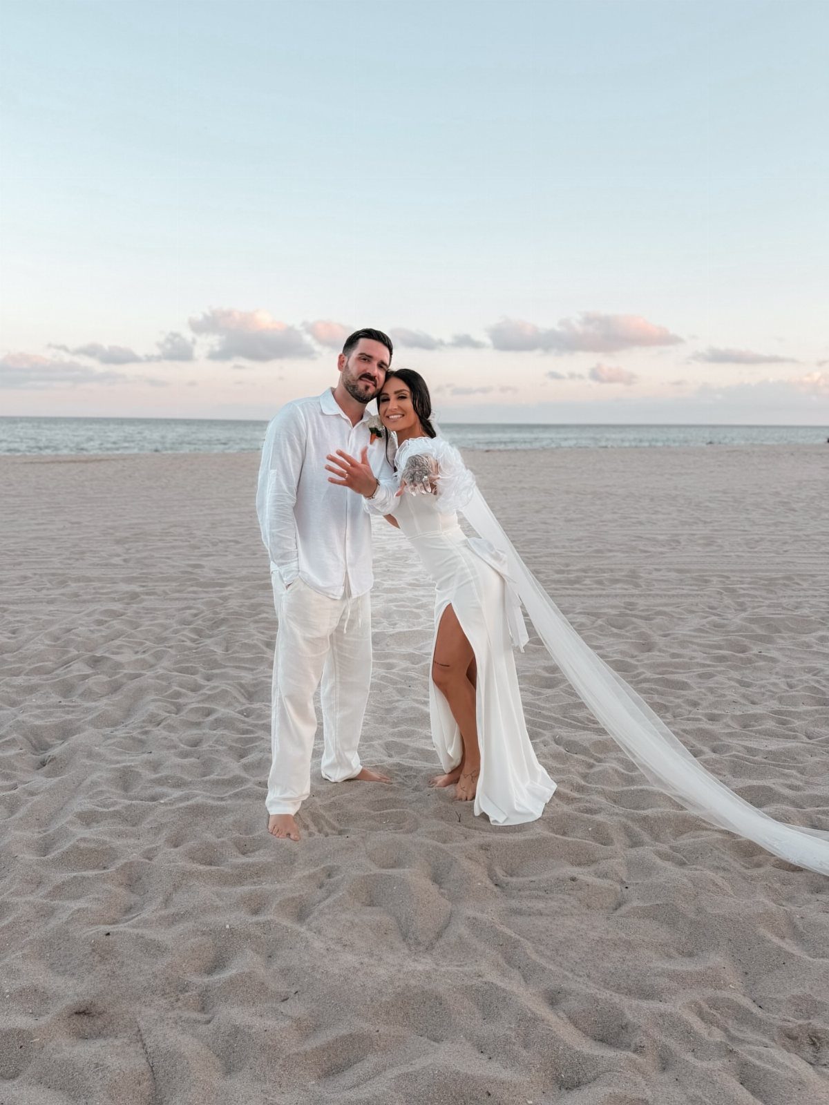 18 Wedding Outfit Theme Ideas for 2026: Beach, Boho, Gatsby & More 2 A bride and groom posing together on a sandy beach during sunset.