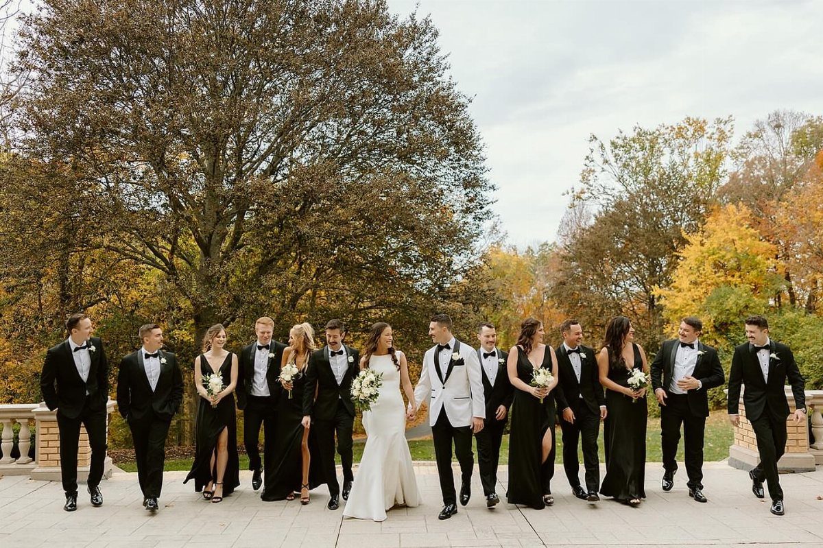 18 Wedding Outfit Theme Ideas for 2026: Beach, Boho, Gatsby & More 11 Wedding party in black and white formal attire walking together outdoors against autumn trees.