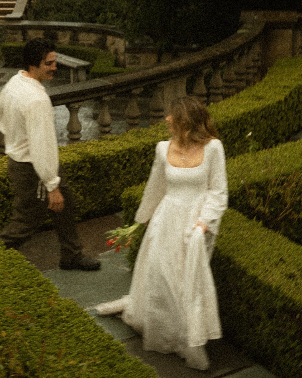 18 Wedding Outfit Theme Ideas for 2026: Beach, Boho, Gatsby & More 12 A woman in a white dress walking through a lush green garden holding red flowers.