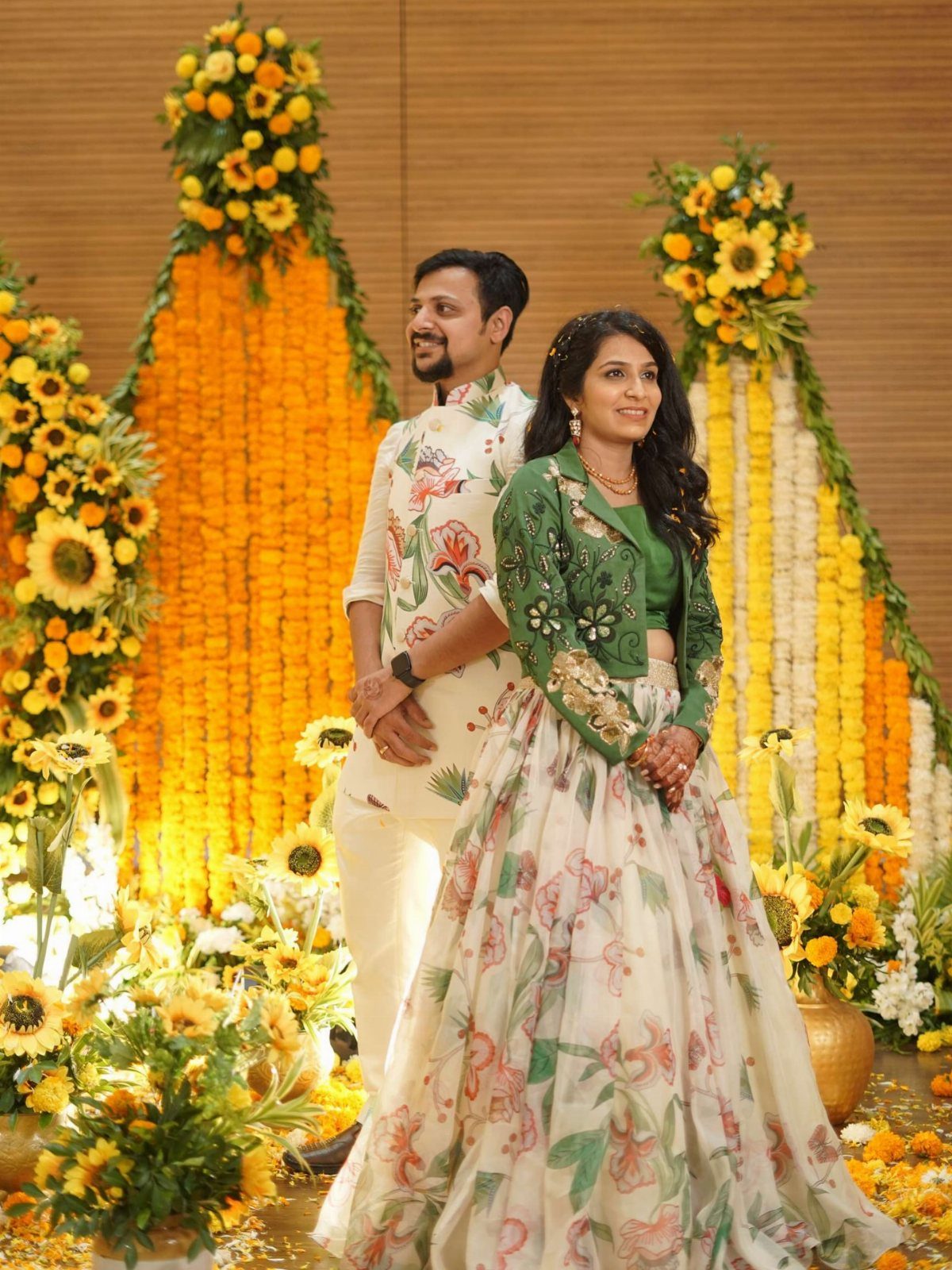 18 Wedding Outfit Theme Ideas for 2026: Beach, Boho, Gatsby & More 18 A couple in floral patterned wedding attire posing before bright yellow flower arrangements.