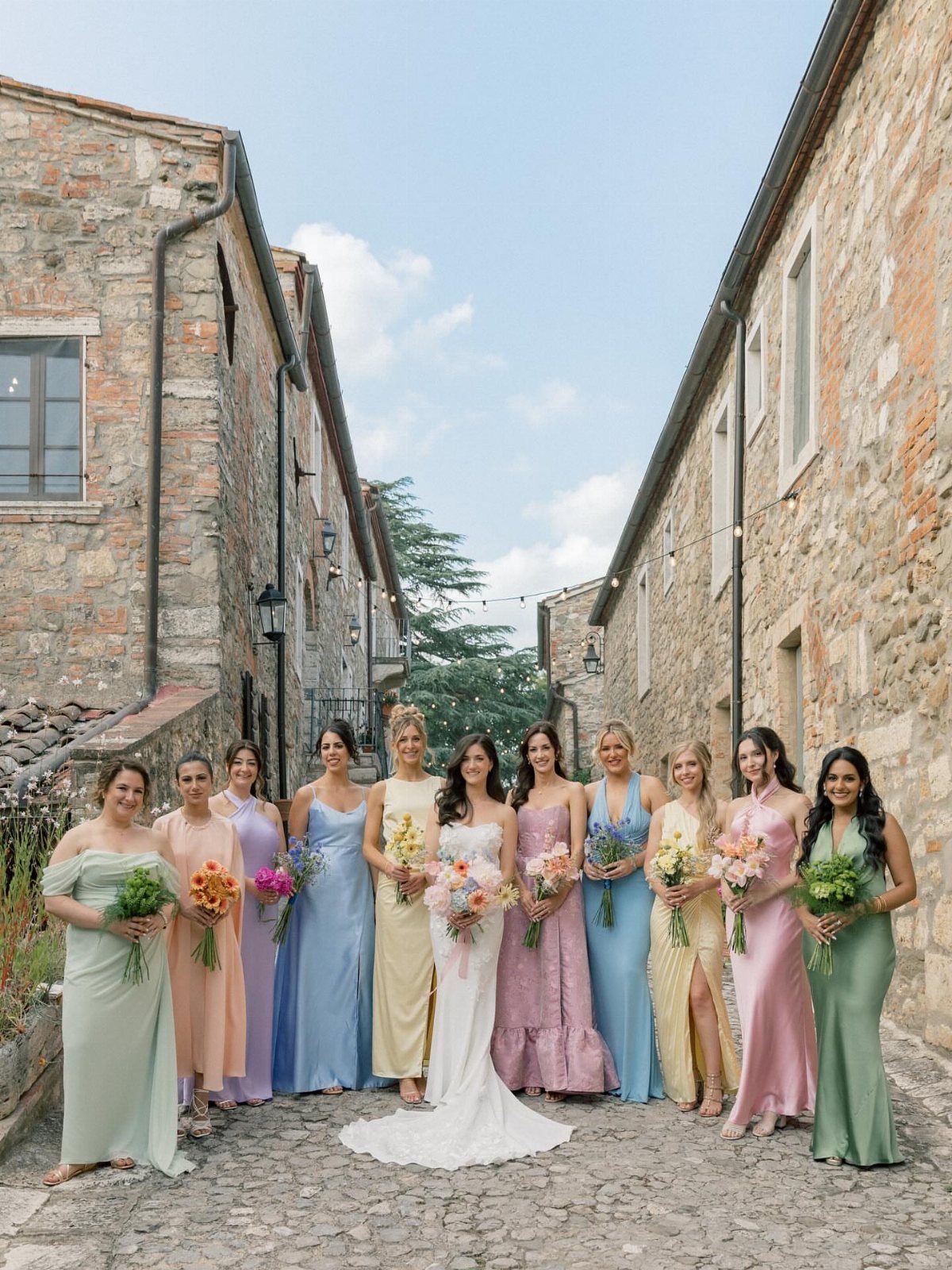 Wedding Party Colors: 13 Real Palette Ideas for Your Bridal Party 6 Bridesmaids wearing various pastel colored dresses standing in a narrow stone alleyway holding bouquets.