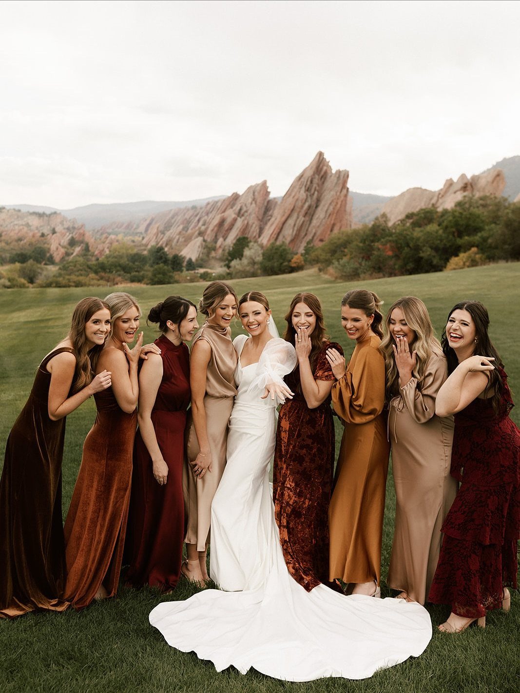 Wedding Party Colors: 13 Real Palette Ideas for Your Bridal Party 2 Bridesmaids in earthy-toned dresses pose with a bride in a grassy field near mountains.