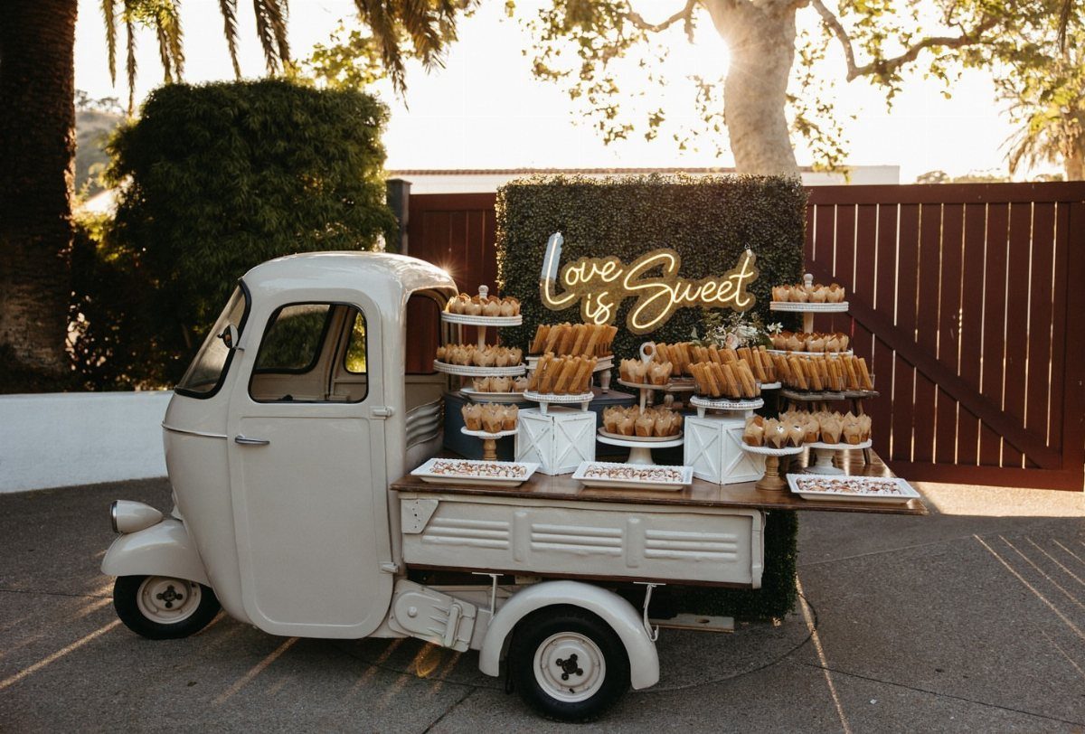 10 Wedding Party Ideas for an Unforgettable Night 7 White vintage dessert truck displaying various pastries and cupcakes under a neon sign.