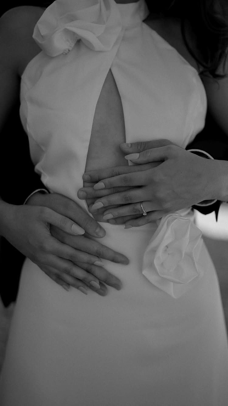 Bride in white dress with groom's hands embracing her waist in black and white.