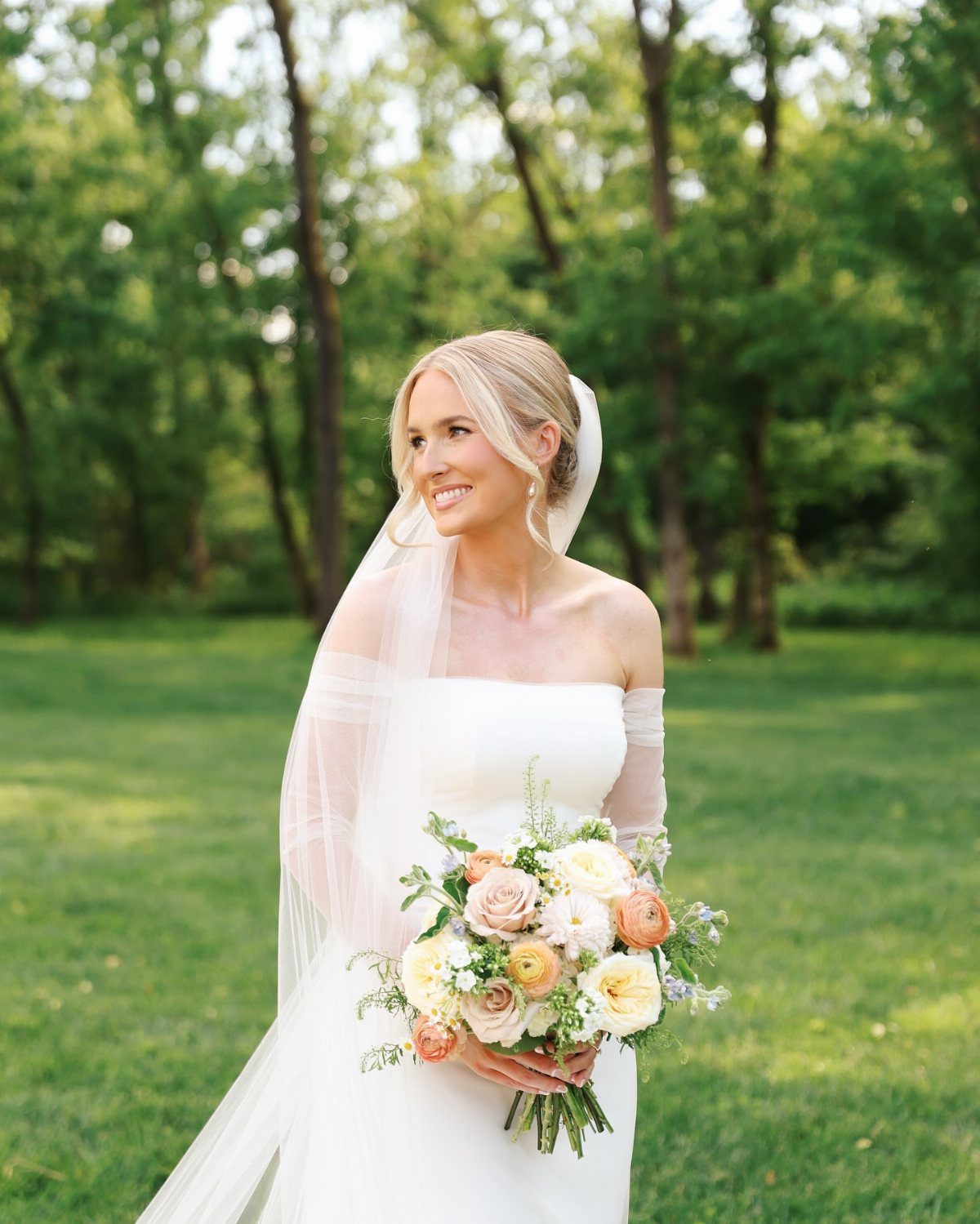 Smiling bride in white dress holding a peach and white floral bouquet outdoors.