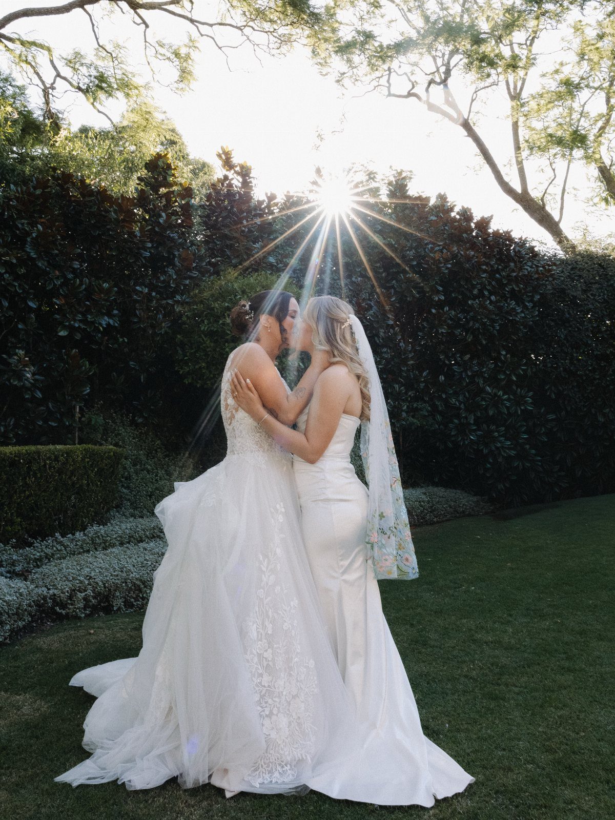 Two brides in white wedding dresses kissing in a sunlit garden