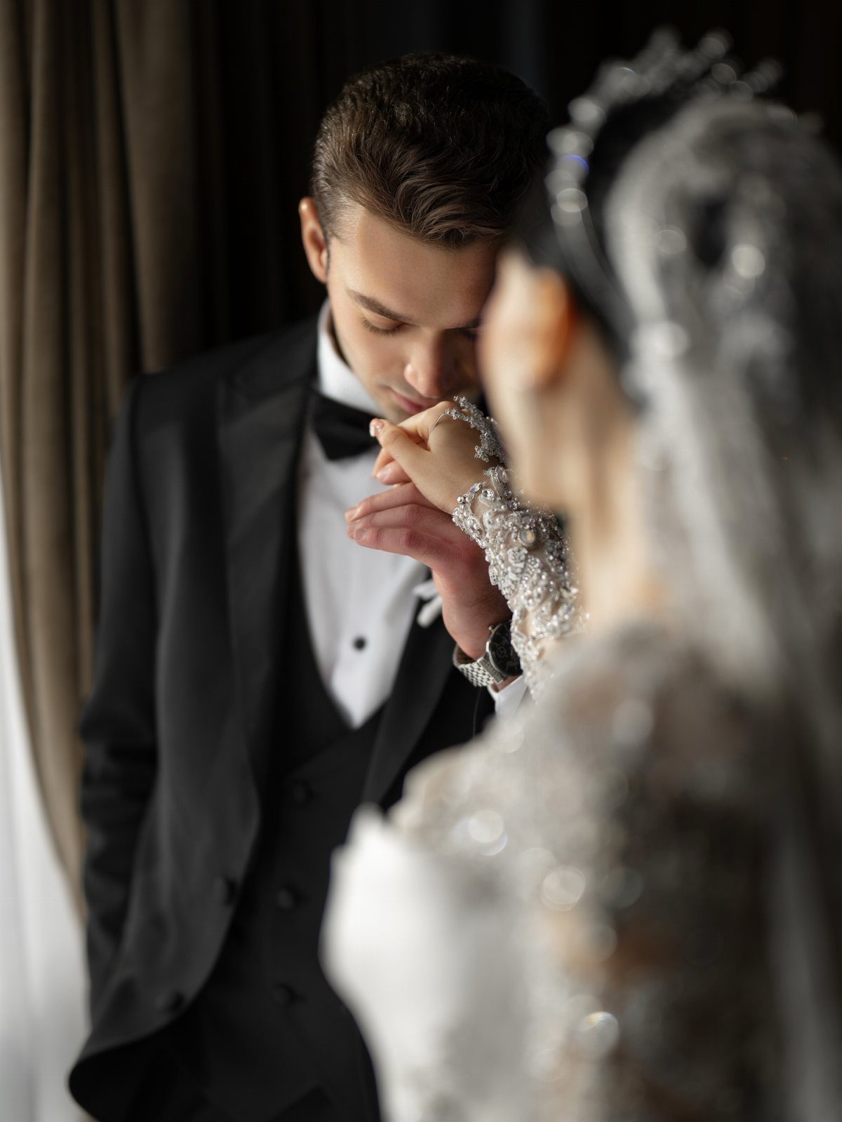 Groom in a black tuxedo kissing the hand of a bride in a beaded gown.