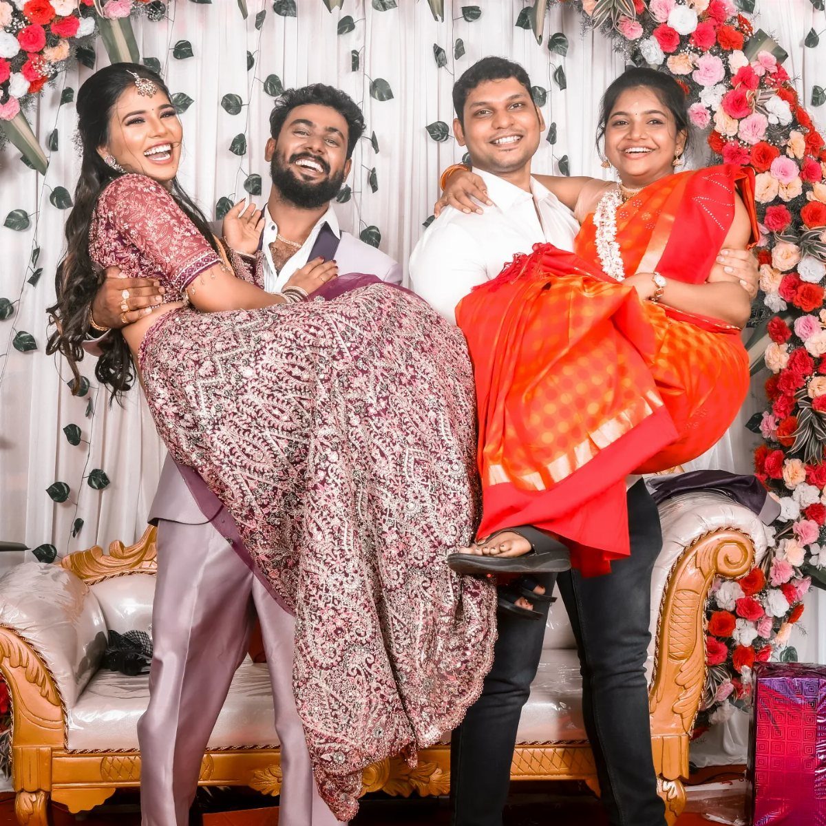 Two men lifting women in traditional Indian sarees, laughing in front of floral backdrop