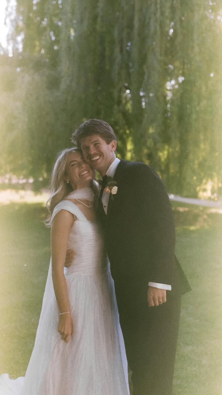 Bride and groom smiling and hugging in a grassy outdoor setting with trees.