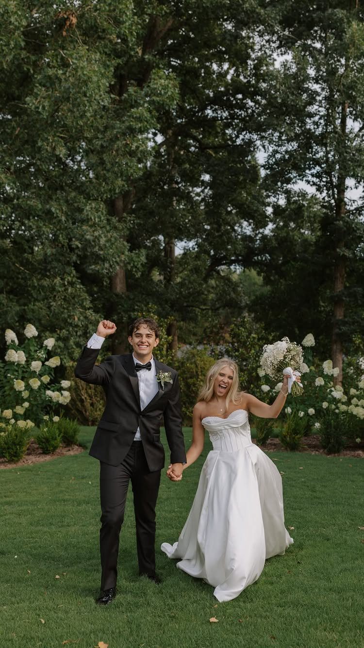Bride and groom cheering and holding hands on a green lawn with trees.