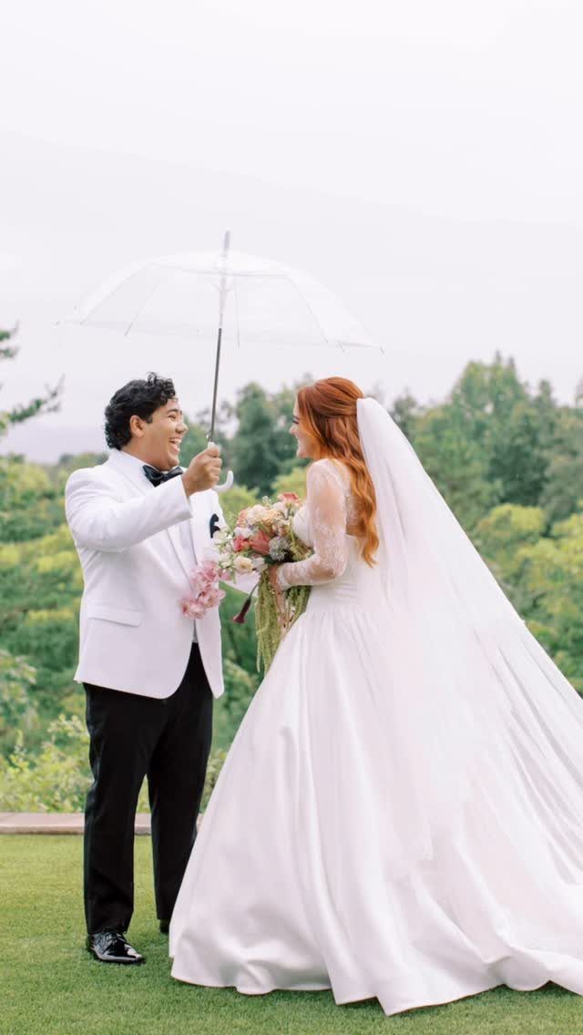 Groom in white tuxedo and bride in white gown facing each other under umbrella.