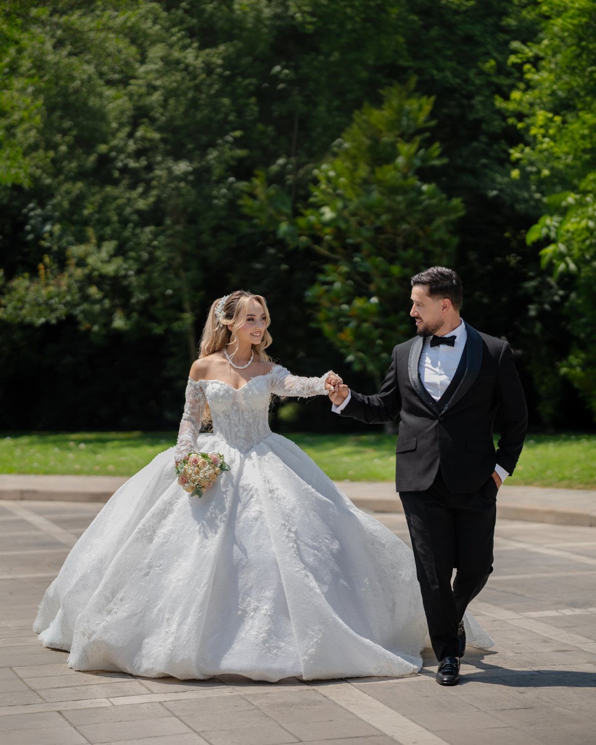 Bride and groom walking hand in hand on a paved path with green trees.