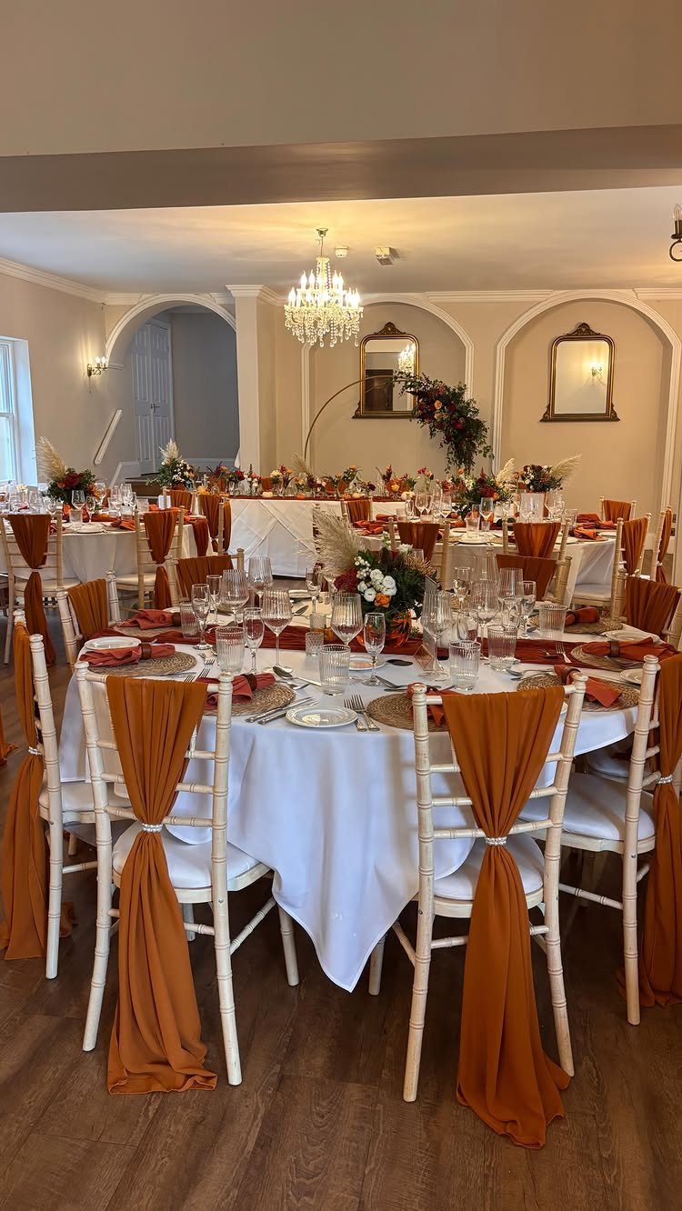 Wedding reception tables with white linens and burnt orange chair sashes and napkins.