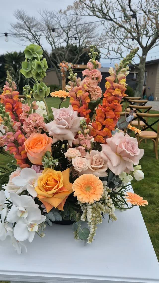 Coral, peach, and yellow floral arrangement with white orchids on a white table.
