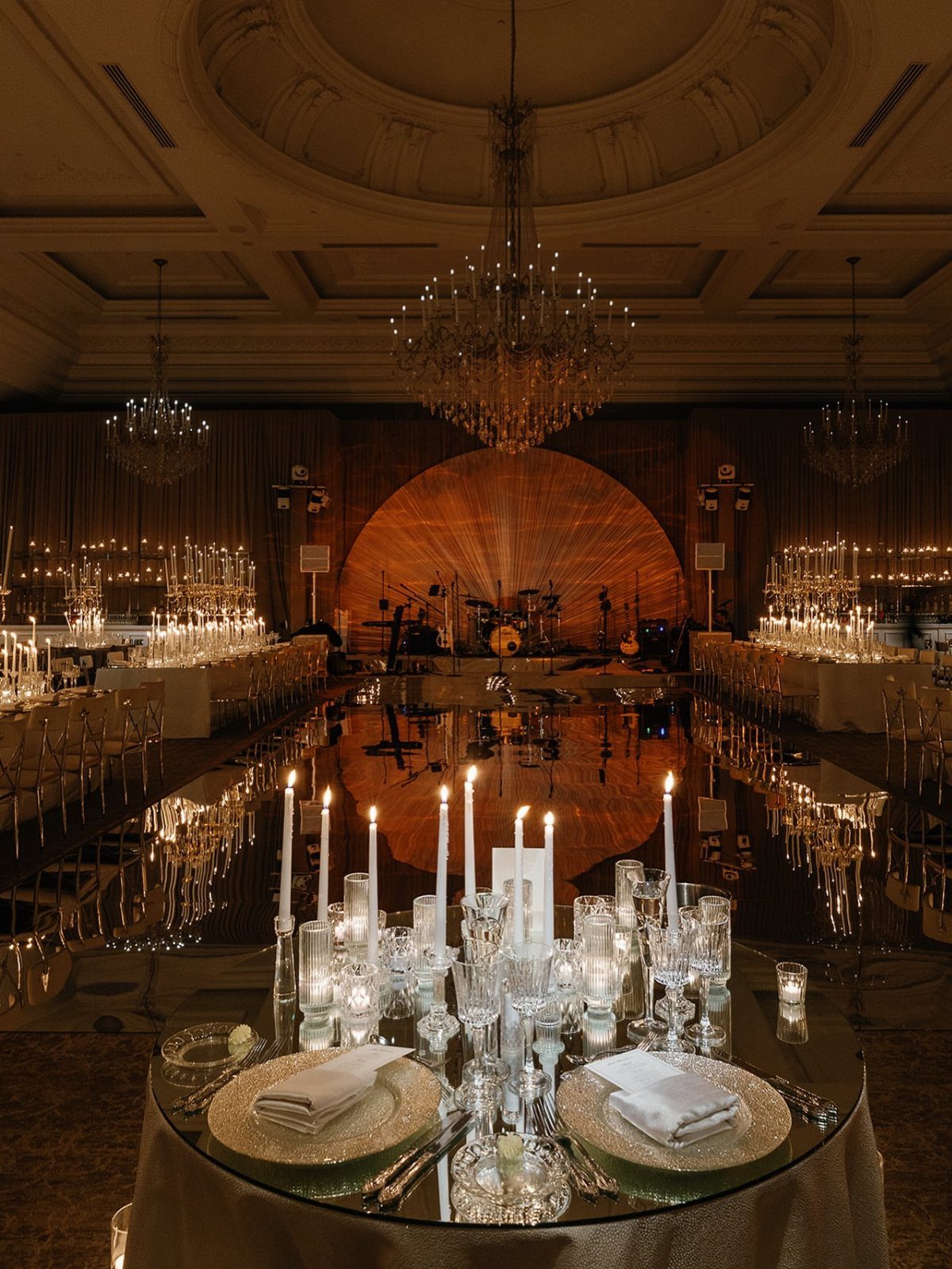 Luxurious wedding reception table with gold plates, white candles, and crystal chandeliers.