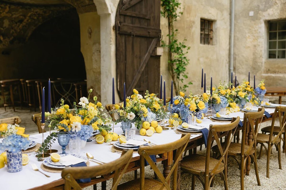 Wedding table setting with yellow flowers, blue candles, and lemons on white linens.