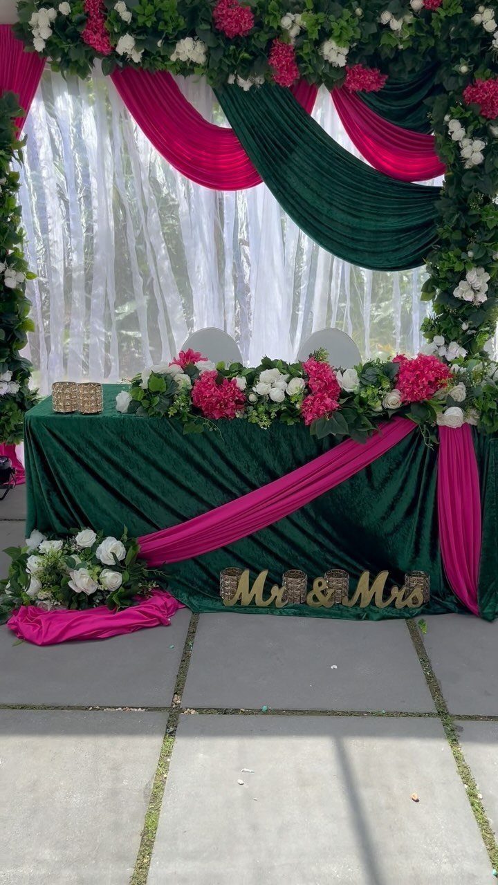 Emerald green and magenta pink draped wedding table with floral arrangements and gold signage.